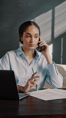 Confident African American multitasking businesswoman in casual clothes, talking on mobile phone with business partners, taking notes while working remotely on laptop from a modern office interior