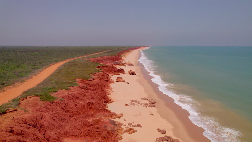 Drone flies forward along James Price Point, revealing dramatic red cliffs, turquoise water, gravel paths, and lush greenery, capturing the rugged and vibrant coastal scenery.