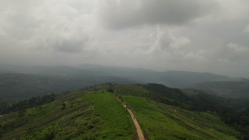 Aerial view of a lone hiker walking on a narrow dirt path along a green mountain ridge under a dramatic cloudy sky. Scenic trekking trail landscape in the tropical highlands with misty background.