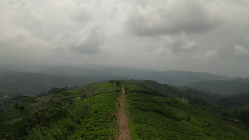 Aerial view of a lone hiker walking on a narrow dirt path along a green mountain ridge under a dramatic cloudy sky. Scenic trekking trail landscape in the tropical highlands with misty background.