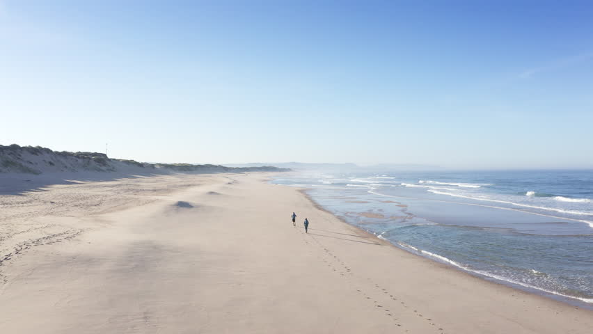 4K aerial view of couple walking by wide sandy beach washed by Atlantic Ocean waves in Portugal casting long shadows in low sunrise light. Shot during the Camino Portuguese —  popular pilgrimage route