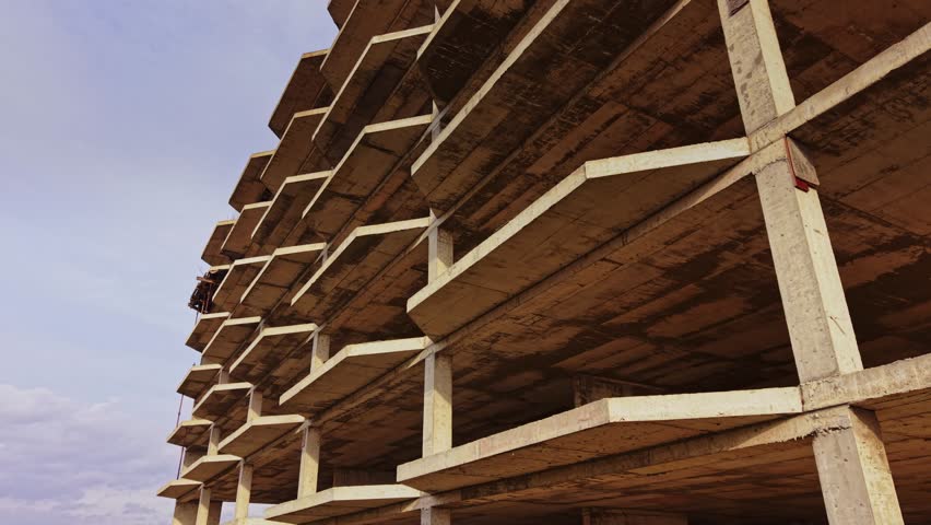 Aerial view of a large unfinished concrete building showing multiple levels and exposed beams. Workers are seen engaging in construction activities in the background.