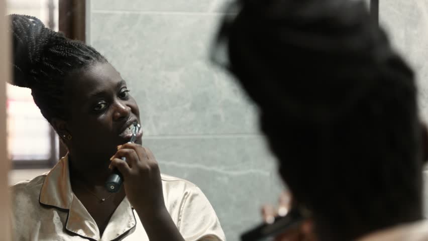 Young African woman in silk pajamas brushes her teeth, reflected in a bathroom mirror, daily self-care and morning routine