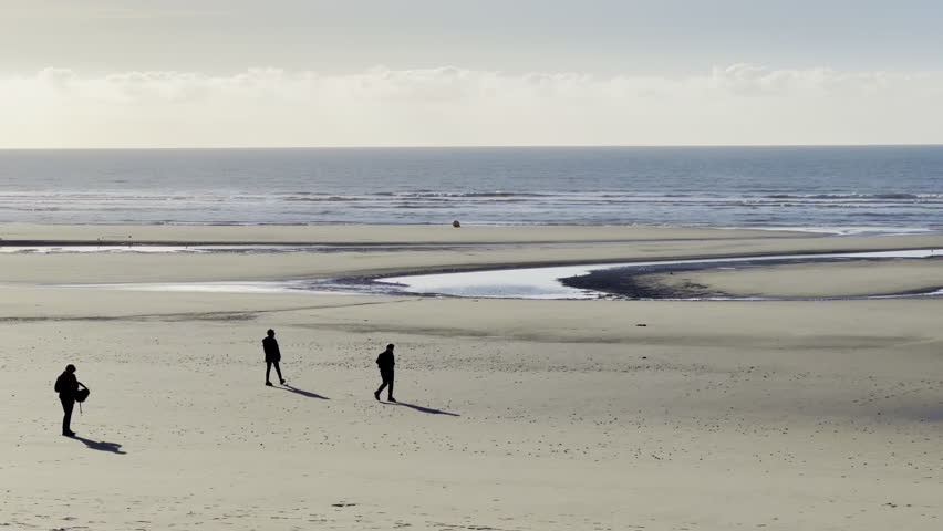 Wide sandy beach at low tide with walking silhouettes and bright winter sun over calm sea at Fort Mahon Quend northern France, panoramic empty shoreline with blue sky, tidal pools and long shadows