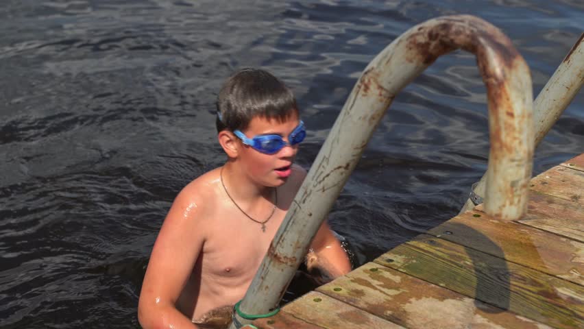 Boy swims in lake and climbs out at a public dock in summer sun