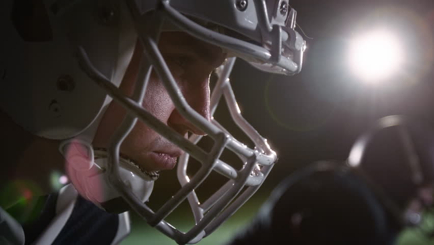 Side close-up of face of young Caucasian male gridiron football player, wearing protective helmet, staring at rival team during standoff in scrimmage line, parting lips and roaring