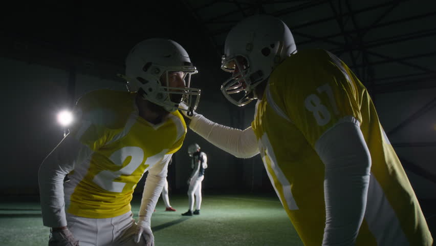 Medium shot of two male gridiron football players in helmets and padded body armor discussing attack or defense strategy, huddling then running off, during training game at dark sports arena