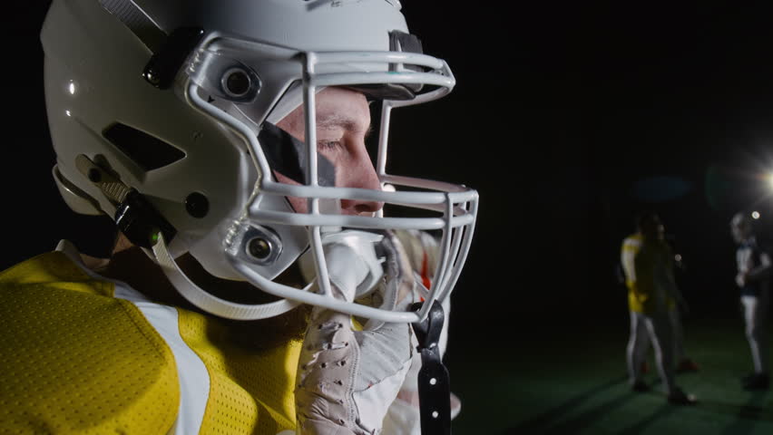 Side close-up of young Caucasian male gridiron football player, dressed in jersey and white gloves, putting on and fastening protective safety helmet while getting ready for training with team