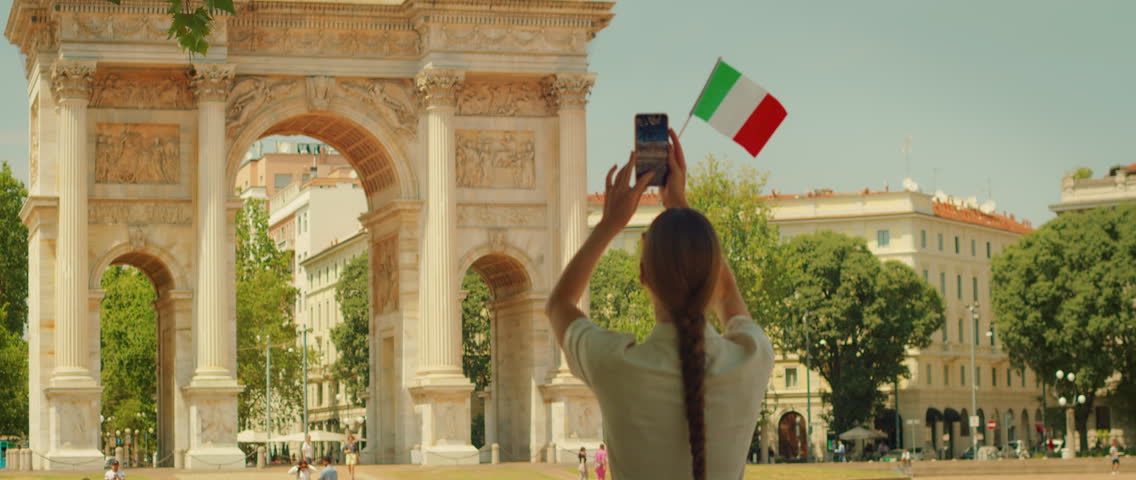 Woman waves Italian flag and takes photo near historic arch in Milan on sunny day