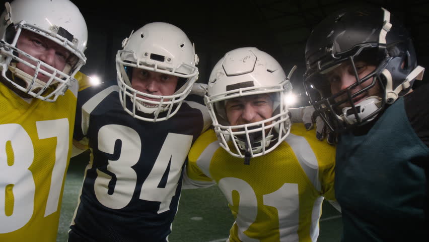 Medium portrait of four happy Caucasian male gridiron football team players standing together on pitch after game, wearing helmets and mouthguards, hugging, smiling and looking at camera