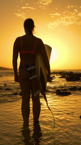 SILHOUETTE, LENS FLARE, VERTICAL: After surfing, a surfer stands ankle deep in the ocean watching waves break at sunset. Moment of reflection by a young woman in bikini after an evening surf session.