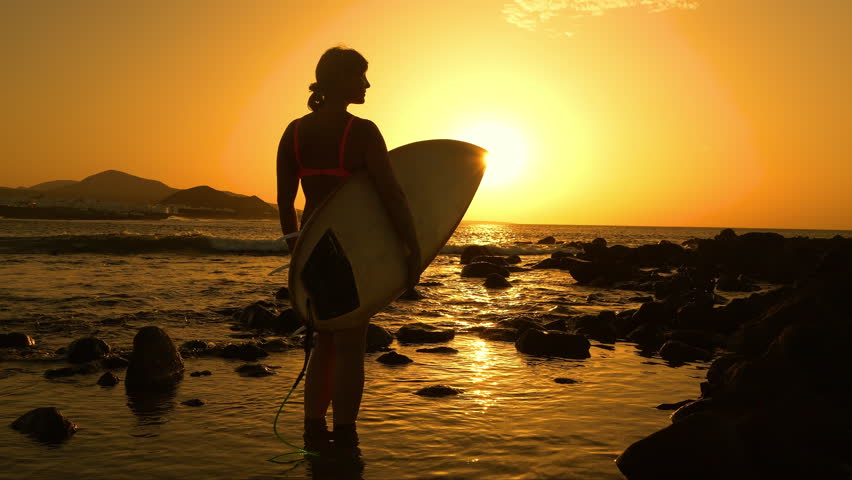 SILHOUETTE, LENS FLARE: Female surfer stands in shallow water, shielding her eyes as she looks out to sea at golden sunset. She is checking out the surf spot before heading for an evening session.