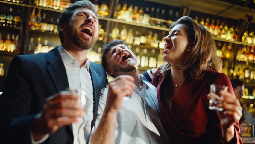 Drunk coworkers singing in bar holding shots in golden light place closeup. Cheerful friends having fun on corporate party meeting at karaoke. Three business people celebrating holiday in night cafe
