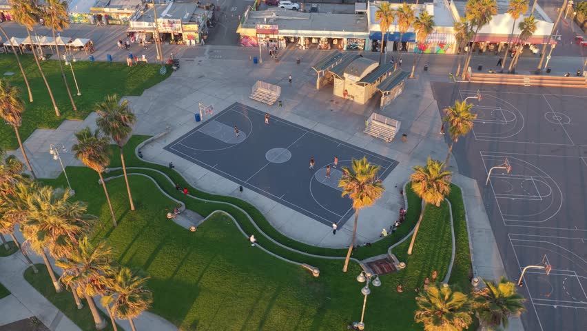Aerial view of Venice Beach basketball courts surrounded by palm trees, skate culture, and colorful storefronts during golden hour in Los Angeles.