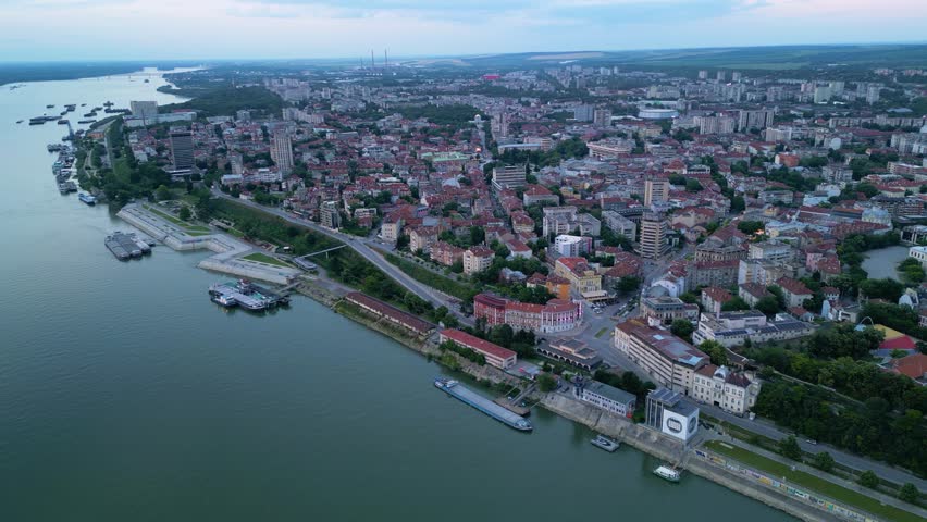 Aerial drone view of historic riverside city with dense red-roofed buildings, waterfront promenade, moored ships, and distant industrial skyline along wide curving river at dusk