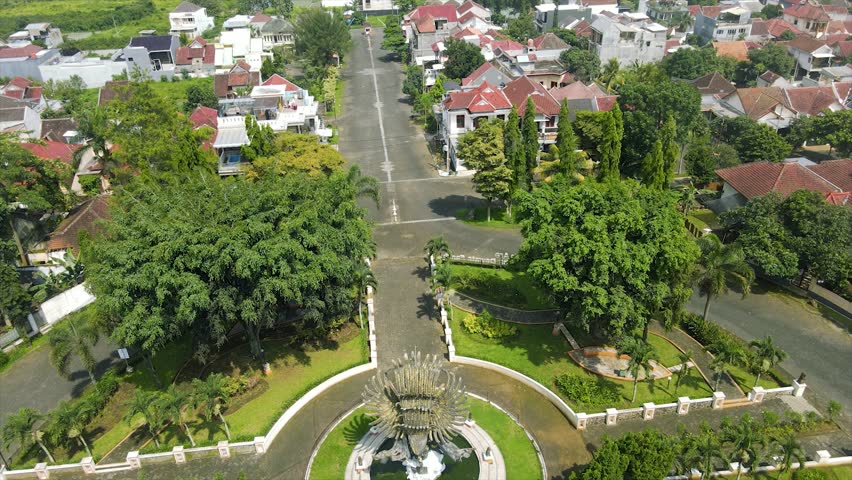 This stunning high-angle aerial photograph captures a grand Garuda monument as the centerpiece of a beautifully landscaped park in a tropical residential area. The golden-hued, multi-feathered statue stands majestically, surrounded by a circular path and manicured green lawns. Tall evergreen cypress trees and vibrant palm trees frame the monument, creating a peaceful and prestigious atmosphere. Beyond the immediate park, the image reveals a clean suburban neighborhood with white-walled houses and tiled roofs, typical of modern Indonesian or Southeast Asian residential developments. In the far distance, a vast city landscape stretches toward the horizon under a bright, partially cloudy sky, showcasing the transition from quiet suburban living to a bustling urban center. The bright daylight emphasizes the lushness of the greenery and the intricate details of the park