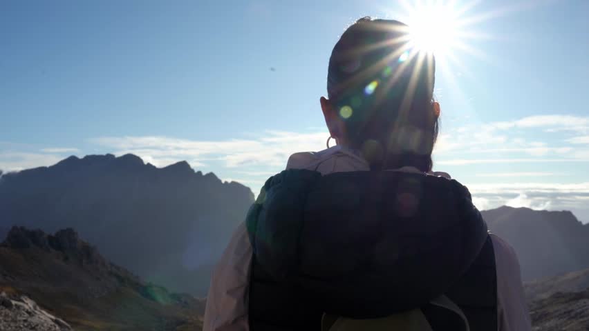 Young woman with a backpack gazes across sunlit mountain peaks and valleys, lens flare adding warmth to this serene hiking scene of solitude, freedom and adventure