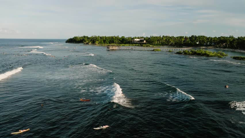Ocean surf with surfers riding waves near wooden pier and tropical shoreline. Cloud 9 Surfing Area. Siargao, Philippines.