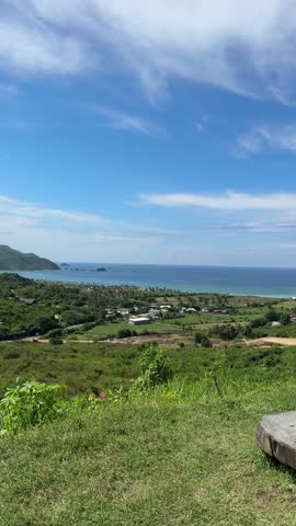 A wooden stump sits on a grassy hill overlooking the ocean and a partly cloudy blue sky under bright sunlight.   