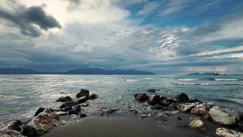 Coastal landscape with rocks, rolling waves and dramatic cloud formations over the sea, static wide shot capturing natural movement of water and changing sky.