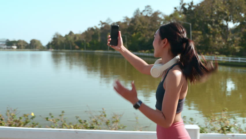 Athletic woman runner using her smartphone for a video call or livestreaming while jogging in a beautiful park with a lake