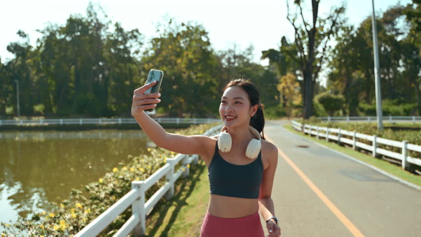 Cheerful Asian runner live streaming or having a video call on her smartphone while jogging. Vlogger creating content for her social media