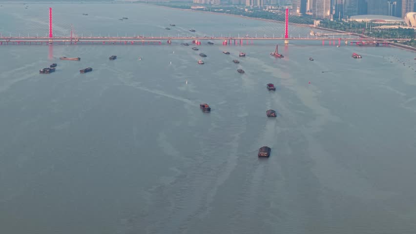 Cargo ships and boats navigate the Qiantang River in Hangzhou China. A red bridge spans the river, with the city skyline visible in the distance