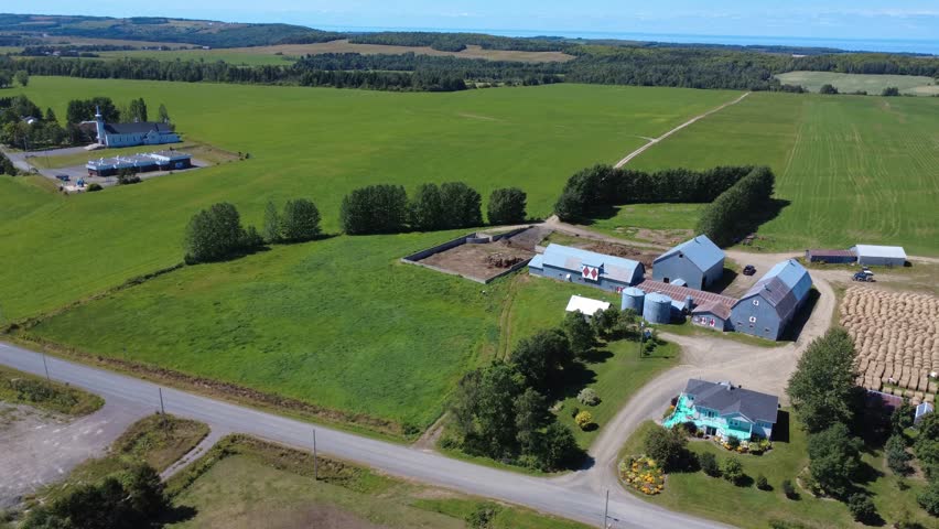 A peaceful rural farm scene with its barns, silos, hay bales, and vast green fields dotted with trees, with a small church in the distance, creates a serene pastoral panorama. Quebec, Canada, 2025.