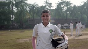 An Indian young, confident girl or female cricket batsman in a jersey and holding a helmet, stands outdoors on the ground, smiles looking at the camera, with teammates preparing in the background - Powered by Shutterstock - Get 15% off with code: PIKWIZARD15