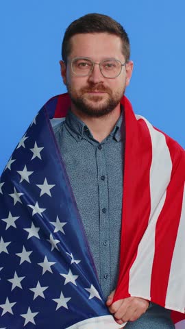 Caucasian middle-aged man in glasses waving and wrapping in American USA flag, celebrating, human rights and freedoms. Independence day. President election. Young adult guy isolated on blue background