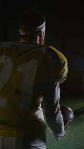 Vertical rear shot of Caucasian male American football player with red beard, in yellow team shirt with 21, padded body armor, standing on pitch before training session, throwing up and catching ball