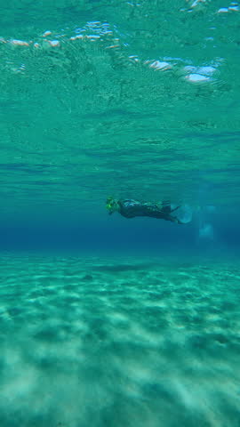 Vertical footage, Underwater shot of Woman in wetsuit swims on surface of turquoise water above sandy bottom on sunny day approaching stairs, takes off fins and climbs up onto pier, Slow motion
