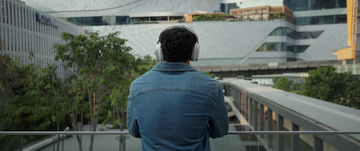Hispanic man wearing denim jacket listens to music via earphones by railing in city anamorphic lens. Latin male with headphone stands on terrace and looks at business center