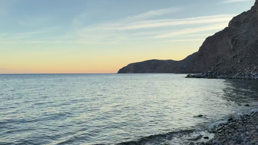 A serene sunset view over the Gulf of Aqaba in Taba, Egypt, where rugged mountains meet calm blue waters. Gentle waves reflect warm evening light beneath a dramatic sky marked by thin cloud streaks left by passing jet aircraft.