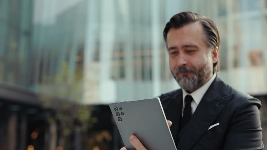 Portrait of grey bearded businessman reads news on digital tablet on downtown backdrop. elderly man entrepreneur scrolls internet via mobile device against business center building