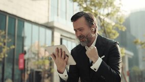 Portrait of senior man supports sports team as watches match via digital tablet on street. Mature businessman expresses excitement of victory using portable computer outdoors - Powered by Shutterstock - Get 15% off with code: PIKWIZARD15