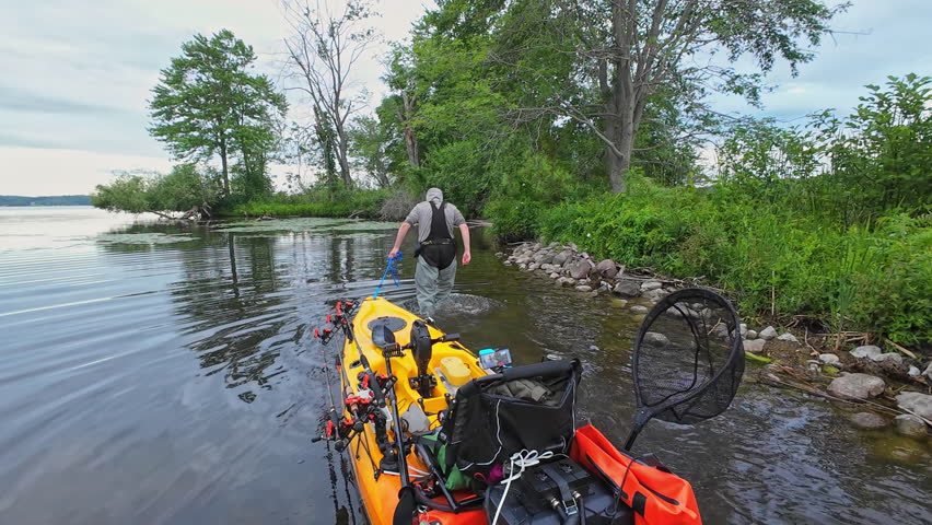 Angler pulling his kayak or water craft to the lake shore for brake, during fishing pov. Fishing pedal kayak in calm lake waters and peaceful landscapes. Water sports bass and pike fishing.