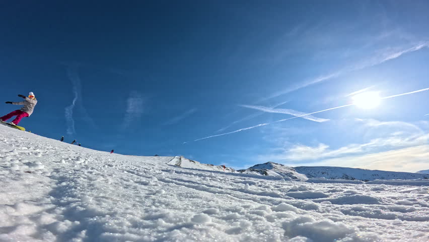 LENS FLARE, LOW ANGLE VIEW, SLOW MOTION: Smiling female snowboarder carving toward camera on snowy piste at ski resort. Snow sprays up around her hand as she leans into turn under bright winter sun.