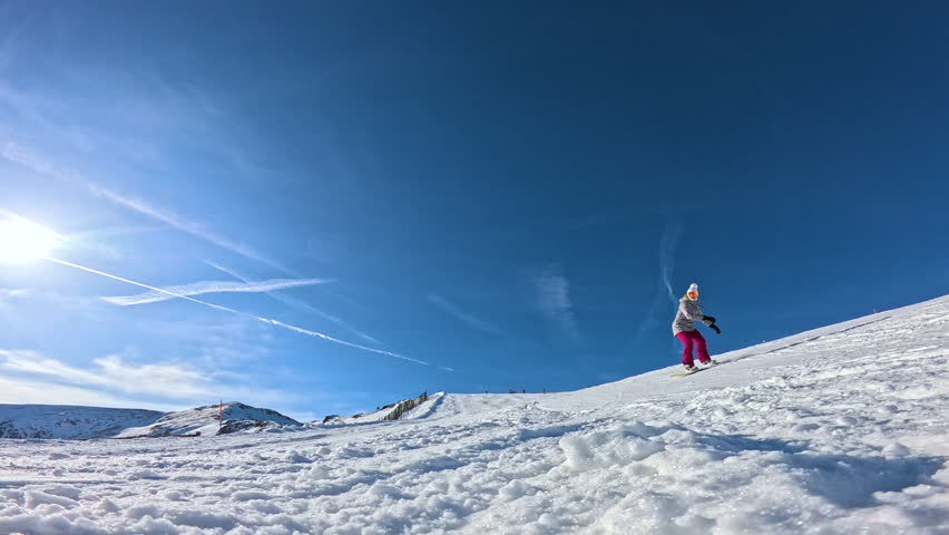 LENS FLARE, LOW ANGLE VIEW, SLOW MOTION: Cheerful female snowboarder drags her hand and sprays snow while she carves across a sunlit slope. Winter fun and authentic snowboard action in Austrian Alps.
