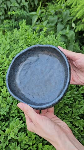 Video shows a handmade ceramic bowl with dark blue matte glaze and uneven organic edges. The object is slowly rotated to demonstrate shape, surface, and handcrafted texture from all sides. Subtle glaze variations and natural imperfections are visible. Neutral background emphasizes artisan pottery, ceramic material, and minimalist tableware design.