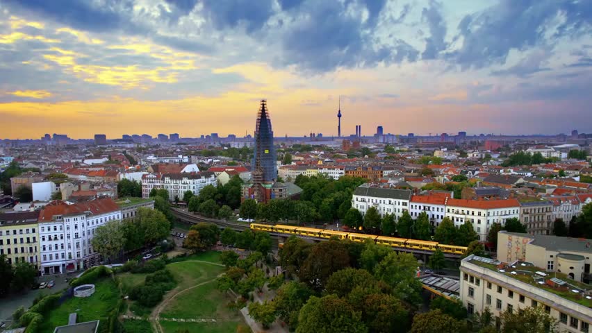 Aerial drone view of Berlin’s Kreuzberg district at sunset, showing residential buildings, lush greenery, moving trains, and the TV Tower in the distance.