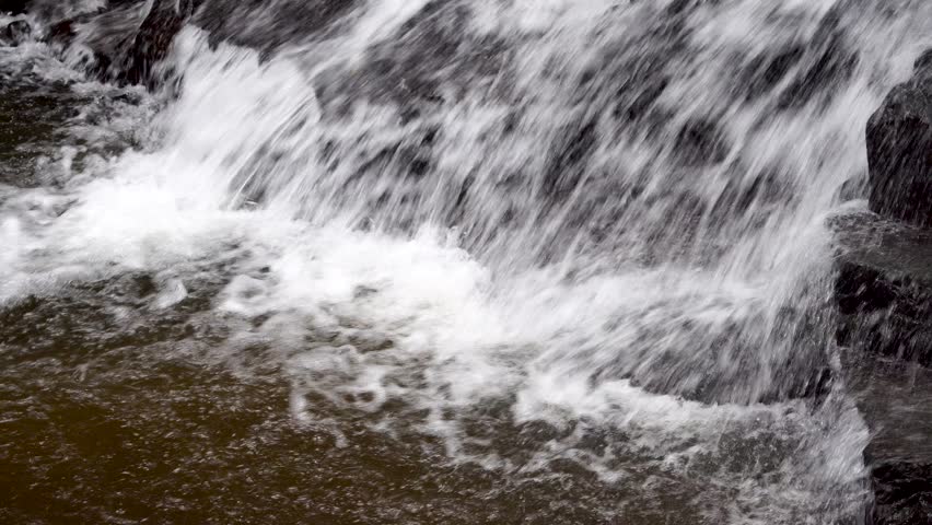 Close-up view of river water cascading over dark rocks, creating white foam and splashes. Turbulent flow of a natural waterfall base with bubbling currents in a stream.