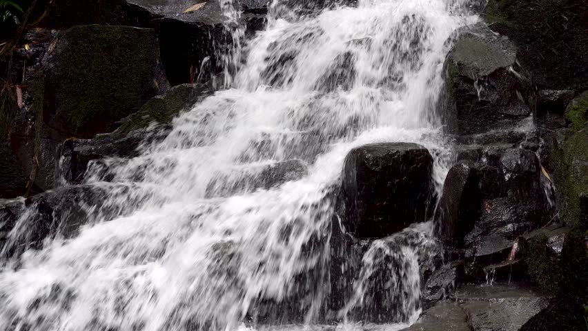 Close up detail of a rushing waterfall cascading over dark rocky cliffs. Fresh white water splashing against wet black boulders in a powerful natural stream flow.