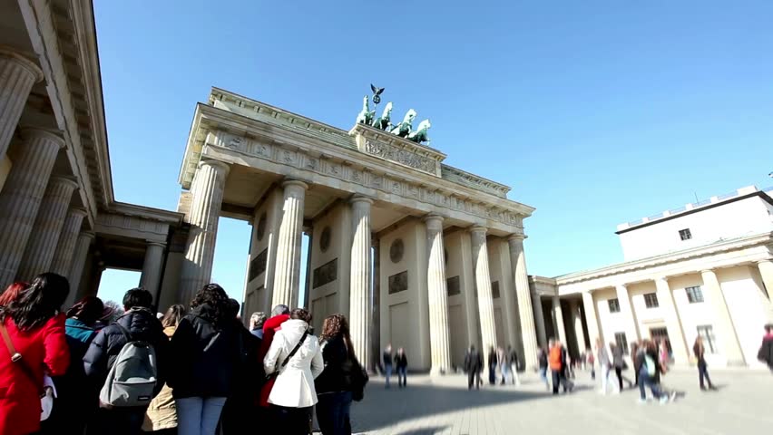 People gather at the iconic Brandenburg Gate in Berlin, enjoying the warm weather and taking in the architectural beauty. The atmosphere is lively with visitors and tourists.