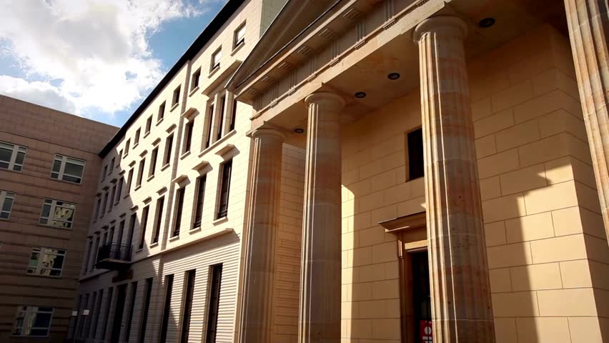 Explore the intricate neoclassical columns of a historic building alongside sleek modern structures, bathed in warm sunlight on a lively city street.