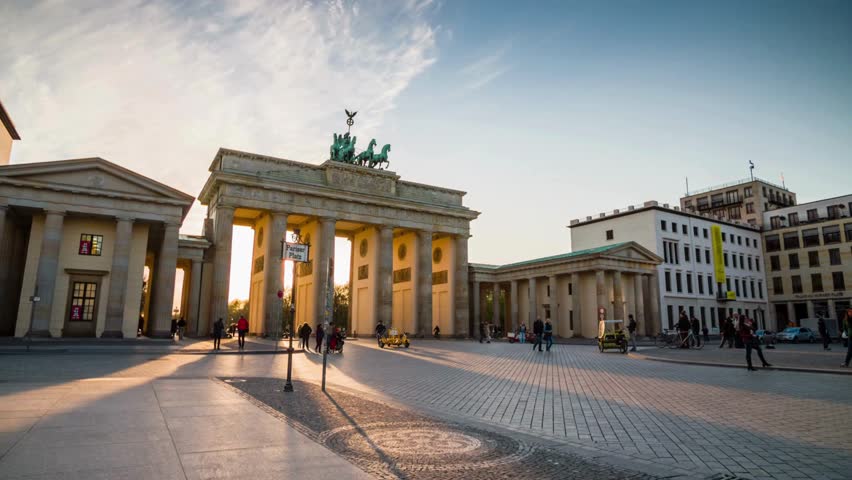 Brandenburg Gate stands majestically at sunset, showcasing vibrant colors against a clear sky. People stroll nearby amid lively urban architecture.