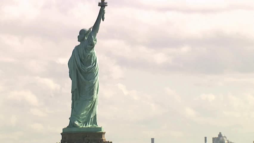 Statue of Liberty stands tall in New York Harbor, showcasing resilience and freedom. Sunlight shines upon this iconic landmark, creating a captivating view. Clouds drift in bright sky.