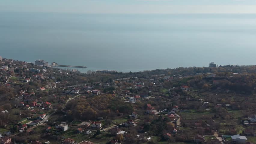 Aerial drone view of coastal city with hillside houses overlooking calm sea, urban landscape meeting nature at seaside during daylight.