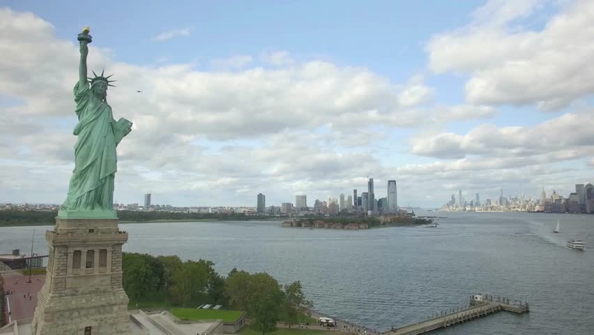 Vibrant skyline stretches across New York Harbor with Statue of Liberty standing proudly. Boats navigate calm waters under fluffy clouds. Perfect setting captures urban life and scenic beauty.