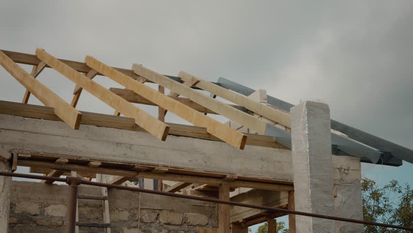 Watch workers install wooden beams on the roof of a house under construction. The structure is taking shape in a peaceful setting, showcasing progress.